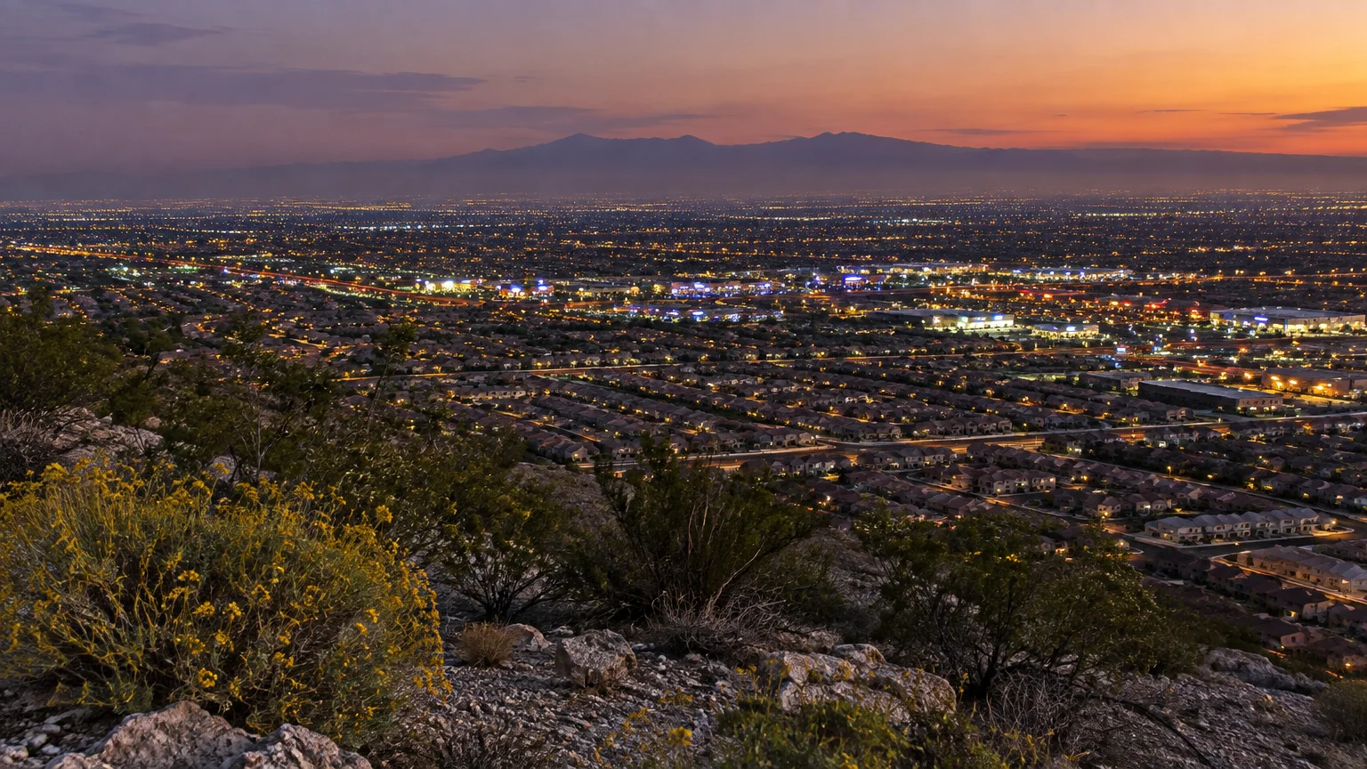 Rocky desert hillside overlooking a sprawling valley of suburban homes and city lights at dusk beneath distant mountains.
