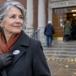 Older woman wearing an I Voted sticker on her coat outside a town hall polling station with people on the steps.