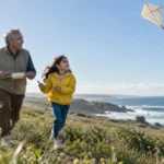 A smiling grandfather and young girl fly a kite while running along a grassy coastal hill overlooking the ocean.