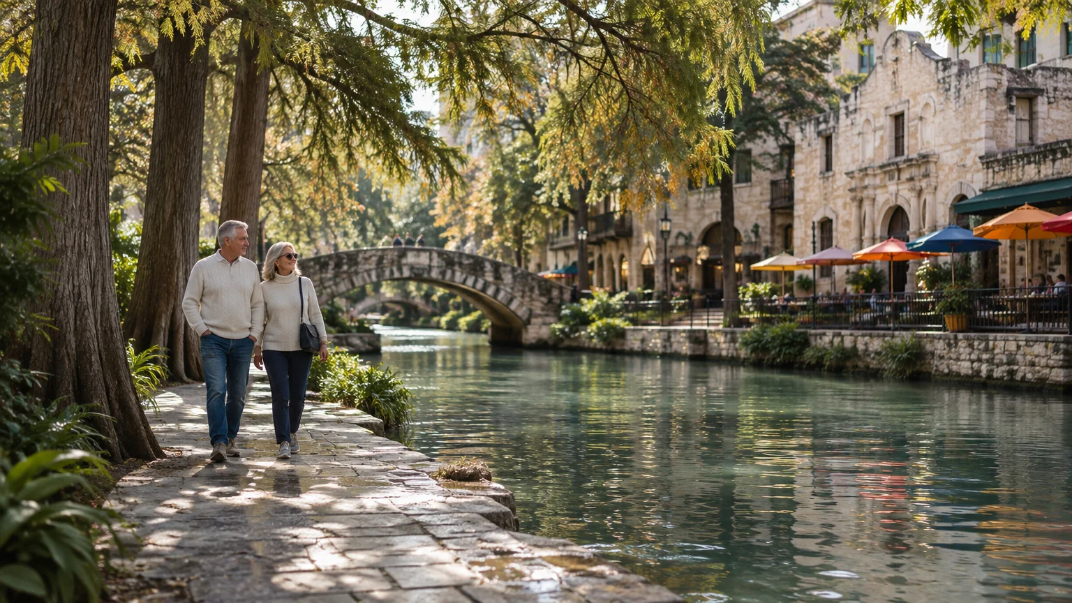 An older couple walks along a stone canal pathway lined with trees, stone bridges, and outdoor restaurant tables.